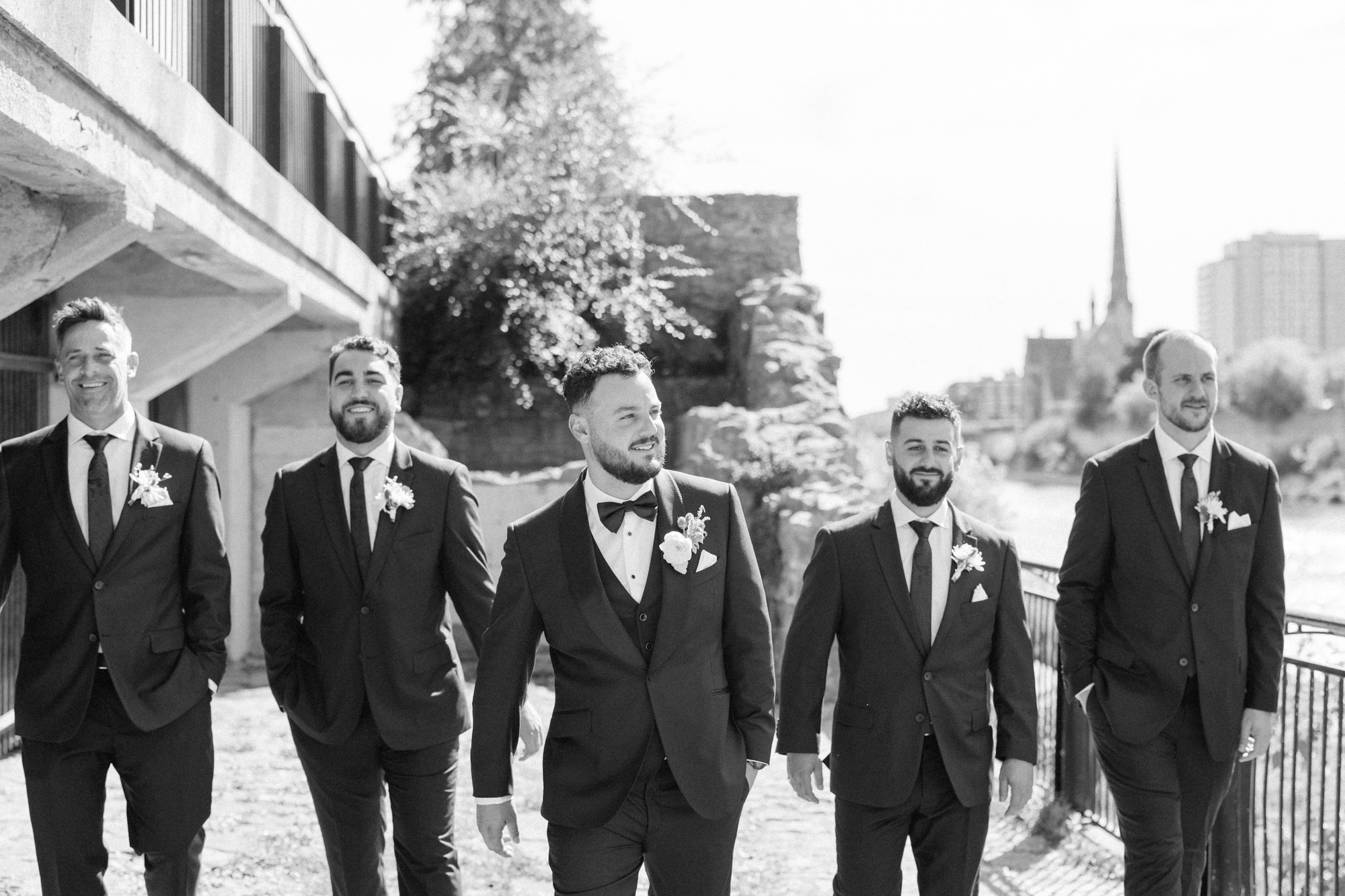 Black and white photo of groom, walking with groomsmen at mill race park in Cambridge